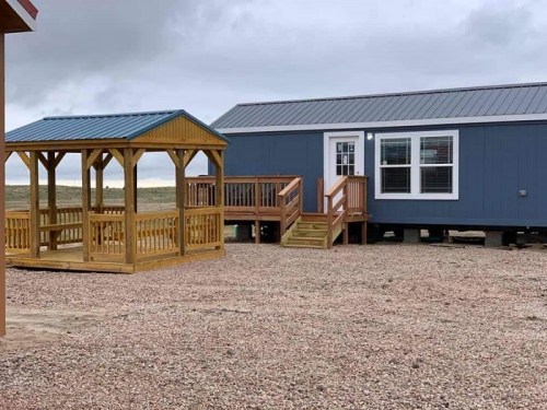 A blue modular home with a wooden gazebo nearby, set on a gravel area under a cloudy sky.