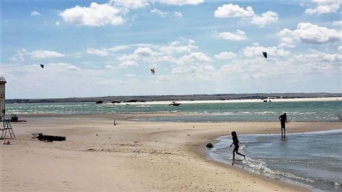 A beach scene with people walking along the shore, kites flying in the sky, and gentle waves lapping at the sand.