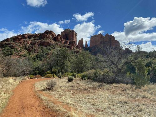 A scenic view of red rock formations under a blue sky with clouds, surrounded by greenery and a dirt path.