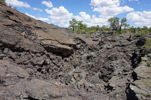 A rugged landscape of dark volcanic rock with scattered trees and a blue sky dotted with clouds.