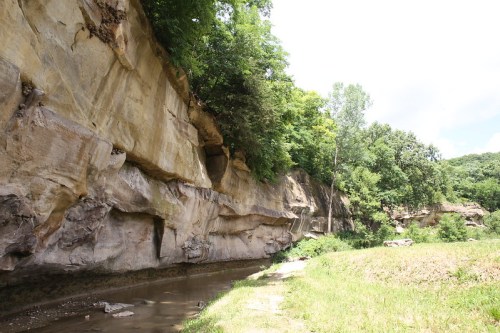 A serene landscape featuring a rocky cliffside with lush greenery and a calm stream running alongside.