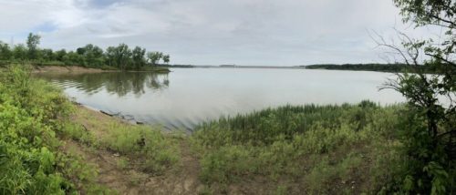 A serene lake view surrounded by greenery under a cloudy sky, with calm waters reflecting the landscape.