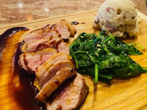 Sliced duck breast with a rich sauce, served with sautéed spinach and a side of creamy mashed potatoes on a wooden board.