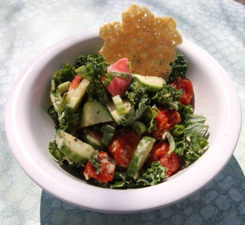 A fresh salad with kale, cucumbers, tomatoes, and a crispy cheese chip on top, served in a white bowl.