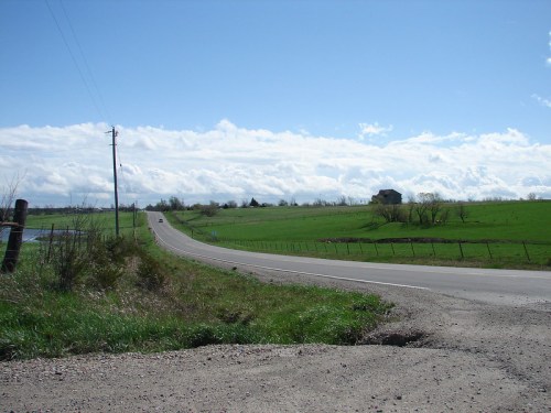A winding road stretches through green fields under a blue sky with fluffy clouds, leading to a distant house.