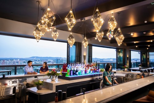 A modern bar with stylish lighting, featuring two people chatting and a bartender preparing drinks against a city view.