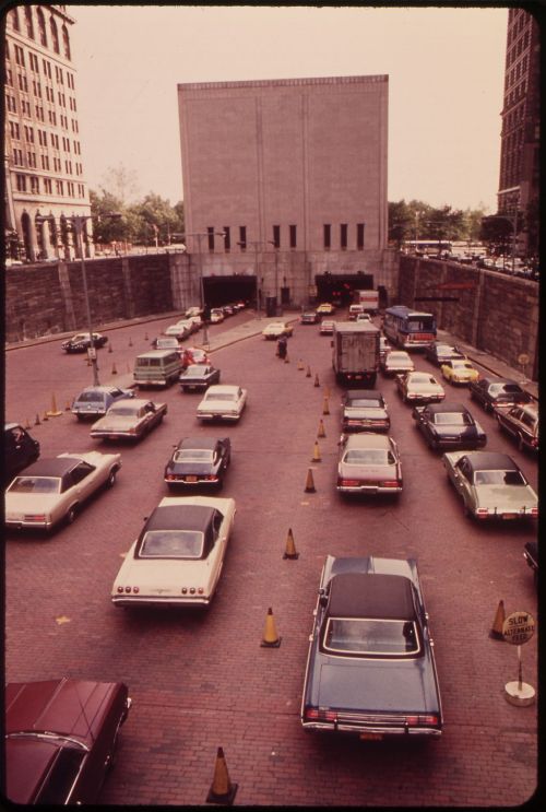A busy parking area with numerous cars and traffic cones, leading to a tunnel entrance in an urban setting.