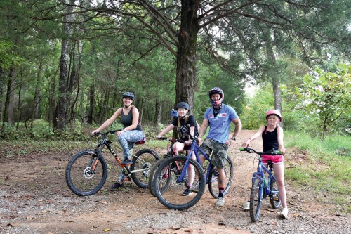 A family of four poses with their bikes on a dirt path surrounded by trees, all wearing helmets and smiling.