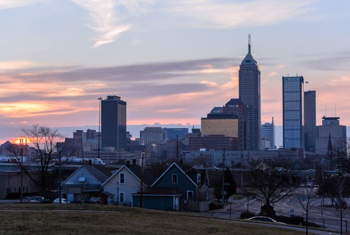 City skyline at sunset, featuring tall buildings and a colorful sky, with houses in the foreground.
