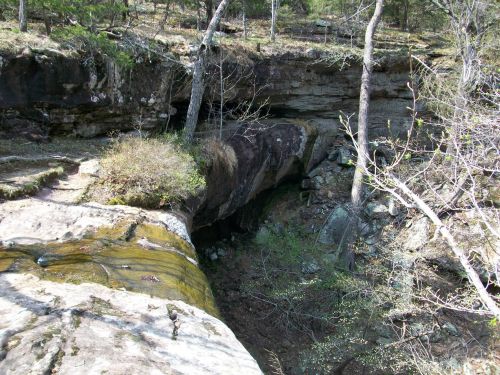 A rocky landscape with a small stream flowing over a ledge, surrounded by trees and greenery.
