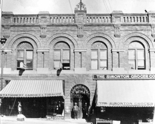 Historic brick building with arched windows, featuring storefronts for a grocer and a stationery shop. Black and white photo.