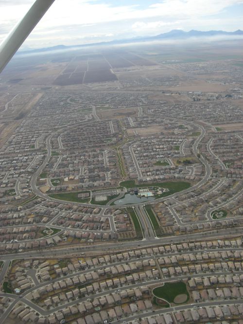 Aerial view of a suburban neighborhood with winding streets and houses, surrounded by open land and distant mountains.