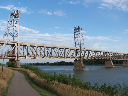 A large metal bridge spans a river, with a pathway alongside and blue skies above. Green vegetation lines the water's edge.