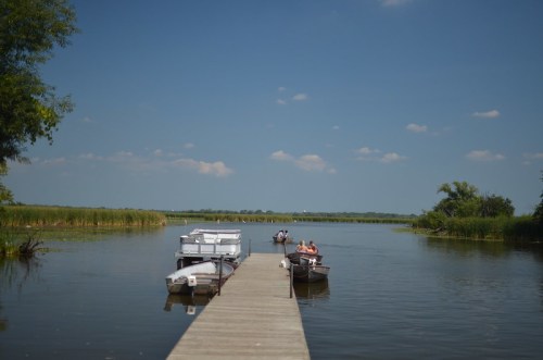 A wooden dock extends into a calm lake, with boats moored and people enjoying the serene landscape under a blue sky.