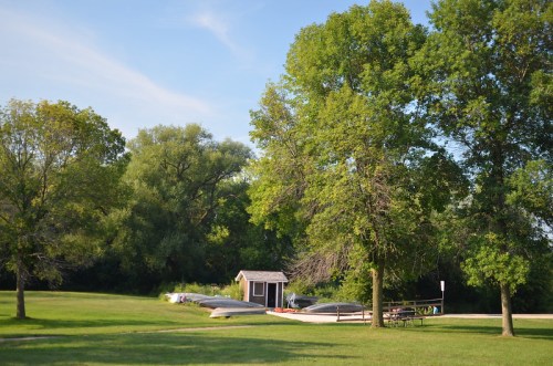 A serene park scene with green trees, a small shed, and canoes resting on the grass under a clear blue sky.