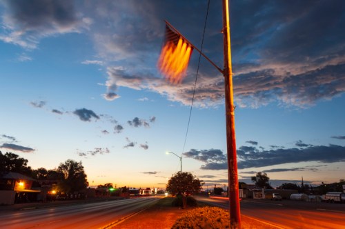 A flag waves on a pole beside a quiet road at dusk, with colorful clouds and a hint of city lights in the background.