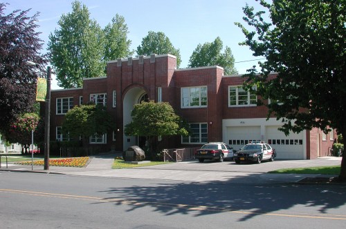 A brick building with large windows, surrounded by trees and parked cars, on a sunny day.