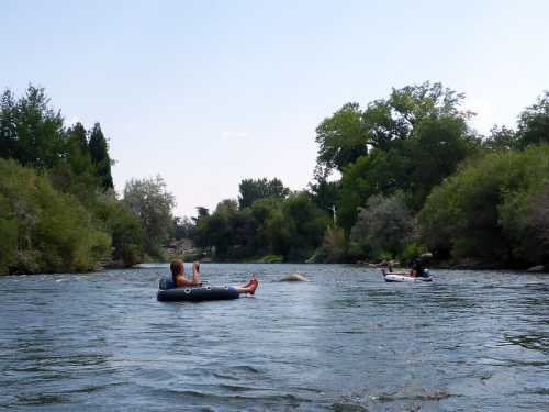 Two people relax on inner tubes in a calm river, surrounded by trees and greenery under a clear blue sky.