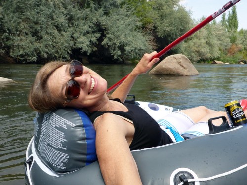 A woman relaxes in a river on an inflatable tube, smiling and holding a paddle, with trees in the background.