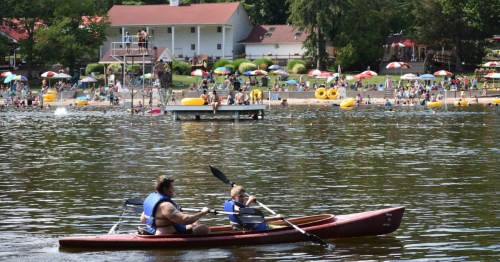 Two kayakers paddle past the busy umbrella-dotted sand beach at Mount Gretna Lake and Beach, Pennsylvania.