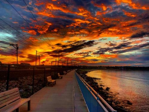 A vibrant sunset over a waterfront path, with colorful clouds and benches lining the walkway beside the water.