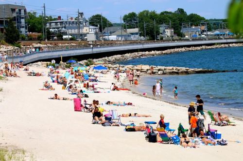 A busy beach scene with people sunbathing, swimming, and relaxing on the sand near the water.