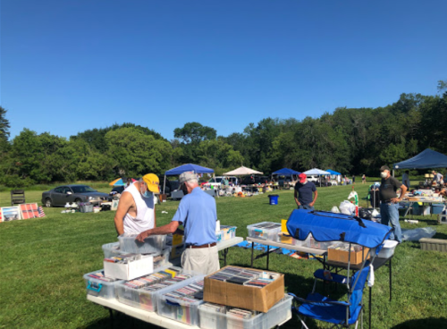Two men interact at a bustling outdoor market with tables of items and tents under a clear blue sky.