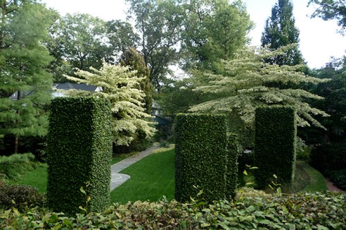 Lush garden with tall, trimmed hedges and trees, featuring a winding stone path and vibrant green grass.