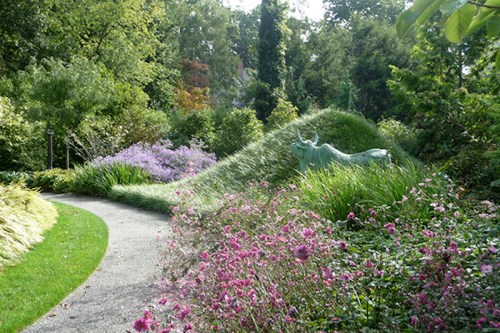A winding path through a lush garden filled with colorful flowers and greenery, featuring a sculpture in the background.