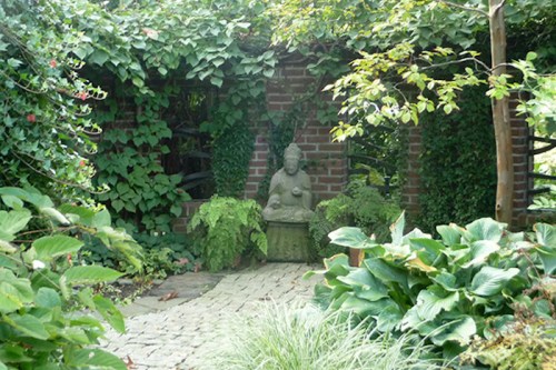 A serene garden scene featuring a stone Buddha statue surrounded by lush greenery and brick walls.