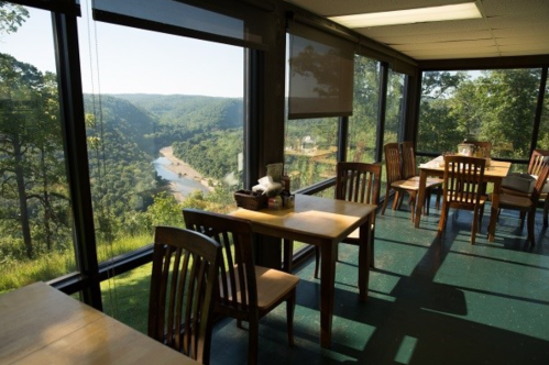 A sunlit dining area with wooden tables and chairs, overlooking a scenic river and lush green hills through large windows.