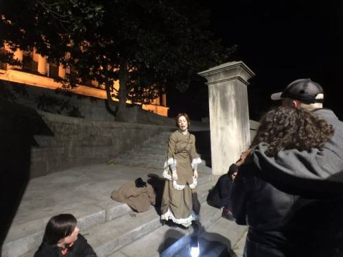 A woman in a vintage dress stands on steps at night, speaking to a small group of people gathered around her.