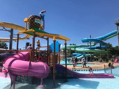 Colorful water park with slides, a play structure, and families enjoying the sunny day by the pool.