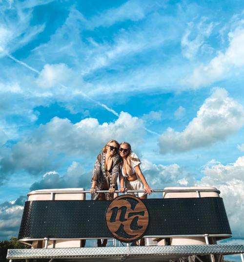 Two women smile and pose on a boat under a bright blue sky with fluffy clouds.