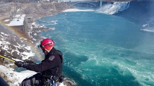 A rescuer in a helmet rappels down a cliff near Niagara Falls, with rushing water and a rainbow in the background.