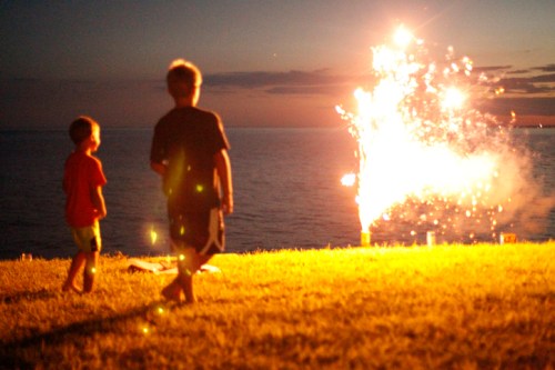 Two children watch fireworks by the water at dusk, with bright sparks illuminating the night sky.
