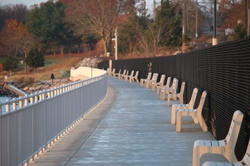 A peaceful walkway lined with benches, surrounded by trees and a black fence, near a body of water at sunset.