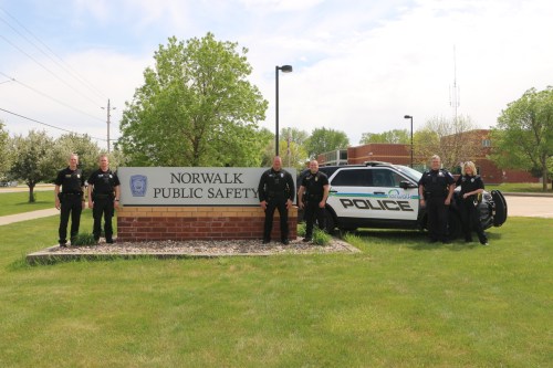 Six police officers stand in front of the Norwalk Public Safety sign and a police vehicle on a sunny day.