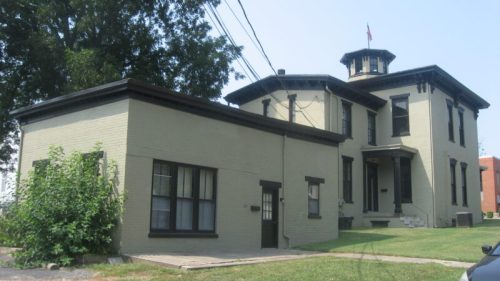 Historic two-story building with a tower, featuring a light-colored brick exterior and a grassy lawn.