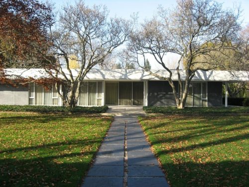 Modern house with large windows, surrounded by trees and a grassy lawn, featuring a stone pathway leading to the entrance.
