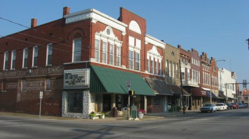 Historic brick buildings line a street, featuring shops and awnings under a clear blue sky.