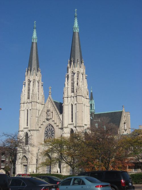 Gothic-style church with two tall spires against a clear blue sky, surrounded by trees and parked cars.