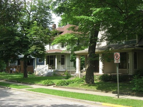 A tree-lined street featuring two large houses with porches and well-maintained lawns.