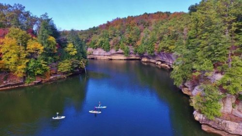 Three paddleboarders on a calm river surrounded by colorful autumn trees and rocky cliffs.