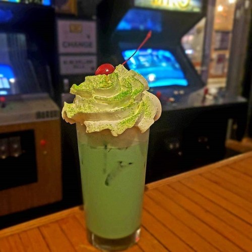 A tall glass of green dessert drink topped with whipped cream and a cherry, set against a retro arcade backdrop.