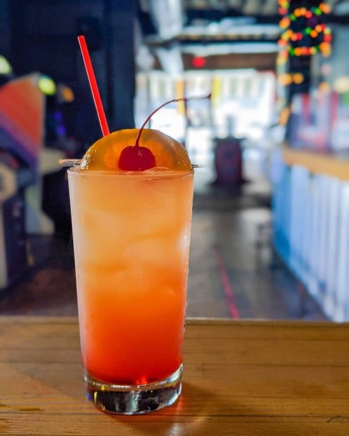 A colorful cocktail with ice, a cherry, and an orange slice, served in a tall glass on a wooden table.