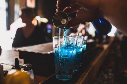 A hand pours a blue liquid into a glass filled with ice, with more glasses in the background at a bar.