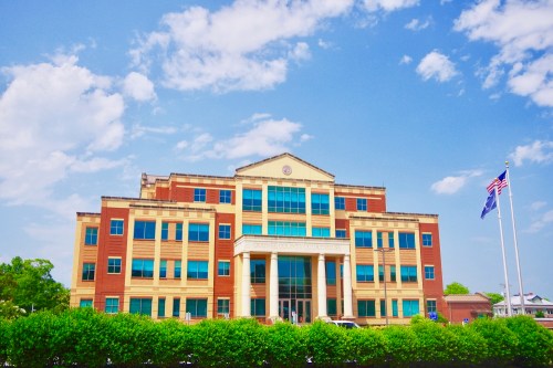 A modern building with large windows, flanked by flags, under a blue sky with scattered clouds. Green bushes in the foreground.