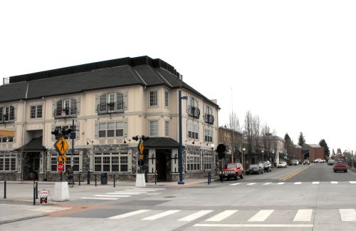A street view of a building with balconies, surrounded by shops and trees, under a cloudy sky.