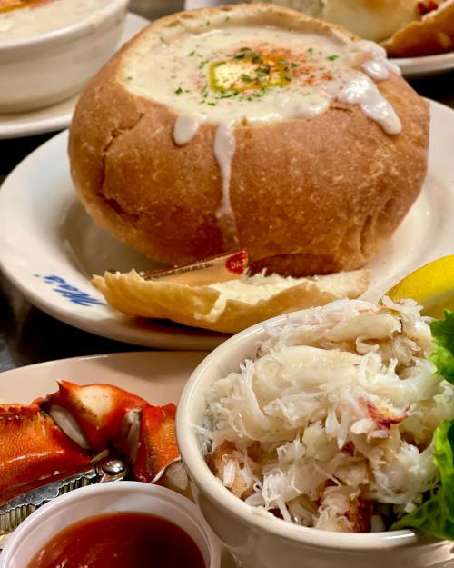 A bread bowl filled with creamy soup, alongside a bowl of crab meat and a lobster claw, garnished with lemon.